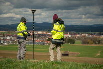 Zwei Personen mit gelben Warnjacken stehen mit technischen Geräten auf einer Wiese. Im Hintergrund ist eine Ortschaft zu sehen.
