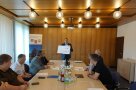 Die Teilnehmenden sitzen an einem langen Tisch im Sitzungssaal. Am Ende steht Michael Neft und hält Urkunden in der Hand.