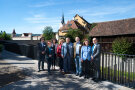 Viele Menschen stehen auf einer Brücke, unter der Wasser durchfließt. Im Hintergrund sind Gebäude, ein Turm und Bäume zu sehen.