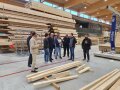 Eine Gruppe Menschen steht in einer Holzfabrik, im Hintergrund ist Holz gelagert.