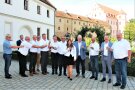 13 Personen stehen im Halbkreis und stoßen mit einem Glas Sekt an.
