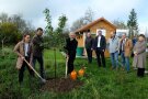 Eine Frau und zwei Männer halten Spaten und einen Obstbaum. Weiter Menschen stehen dabei und sehen ihnen zu.