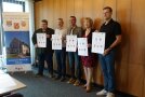 Die Bürgermeisterin und die vier Bürgermeister halten die Urkunde in den Händen und stehen nebeneinander an der Wand.