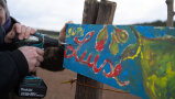 Ein buntbemaltes Holzschild mit der Aufschrift „Luise“ wird an einen Holzpfahl geschraubt.