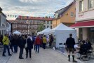 Menschen stehen vor weißen Ständen in einer Stadt. 