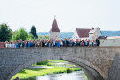 Viele Menschen stehen auf einer Brücke, unter der Wasser durchfließt. Im Hintergrund sind Gebäude, ein Turm und Bäume zu sehen.