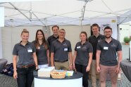 Drei Frauen und vier Männer stehen an einem Stand unter einem Pavillon und lächeln in die Kamera.