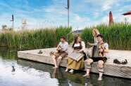 Zwei Frauen und zwei Männer sitzen in Trachtenkleidung mit Musikinstrumenten auf einem Steg und lassen zum Teil die Füße ins Wasser hängen. 