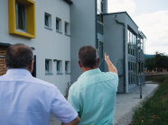 Zwei Männer stehen vor dem Gebäude, mit dem Rücken zur Kamera, ein Mann zeigt auf das Gebäude. 