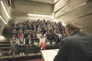 Ein Mann mit dem Rücken zur Kamera spricht zu vielen sitzenden Personen