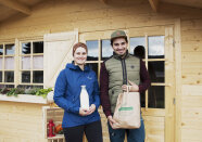 Zwei junge Menschen vor einem Holzhäuschen halten eine Milchflasche und eine Papiereinkaufstüte in den Händen.