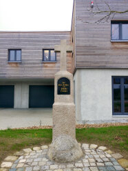 Ein steinernes Kreuz mit der Aufschrift „Zur Ehre Gottes von Familie Fischer“ steht vor einem Gebäude.
