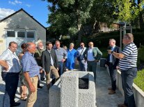 Viele Personen stehen um einen Brunnen und hören einem Mann zu. 