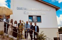 Fünf Männer und zwei Frauen stehen lächelnd an einem Geländer, im Hintergrund ist das neue Dorfgemeinschaftshaus, die Sonne scheint.