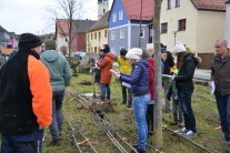 Auf einer Grünfläche stehen viele Menschen, am Boden liegen verschiedene Sträucher und Bäume.