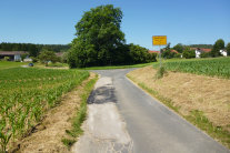 Asphaltierter Weg in schlechtem Zustand, der durch Felder in den Weiler Dettnach führt.