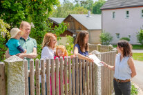 Eine Frau gibt von der Straße aus über den Gartenzaun eine Broschüre an eine Frau und ihre Familie, die im Garten stehen.