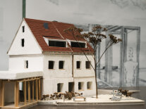 Ein Hausmodell ist in Nahaufnahme zu sehen, es hat drei Stockwerke, eine weiße Fassade und ein rotes Dach.