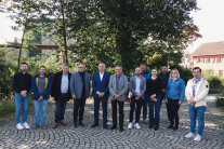 Eine Gruppe von Menschen steht nebeneinander aufgereiht auf einem gepflasterten Platz, im Hintergrund sind Bäume.