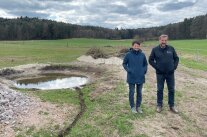Ein Mann und eine Frau stehen auf einem Feld mit einem kleinem Gewässer. 