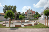 Runde Sitzgelegenheit auf einem Dorfplatz, der durch Bäume, Kirche und Rathaus eingerahmt wird.