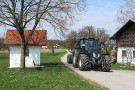 Traktor mit Anhänger fährt auf geteerten landwirtschaftlichen Weg. Am Wegrand steht ein Wohnhaus, gegenüber einer kleinen Kapelle.