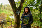Forstministerin Kaniber steht neben einem Baum mit dem Naturwald-Logo und hält ein Tablet in den Händen